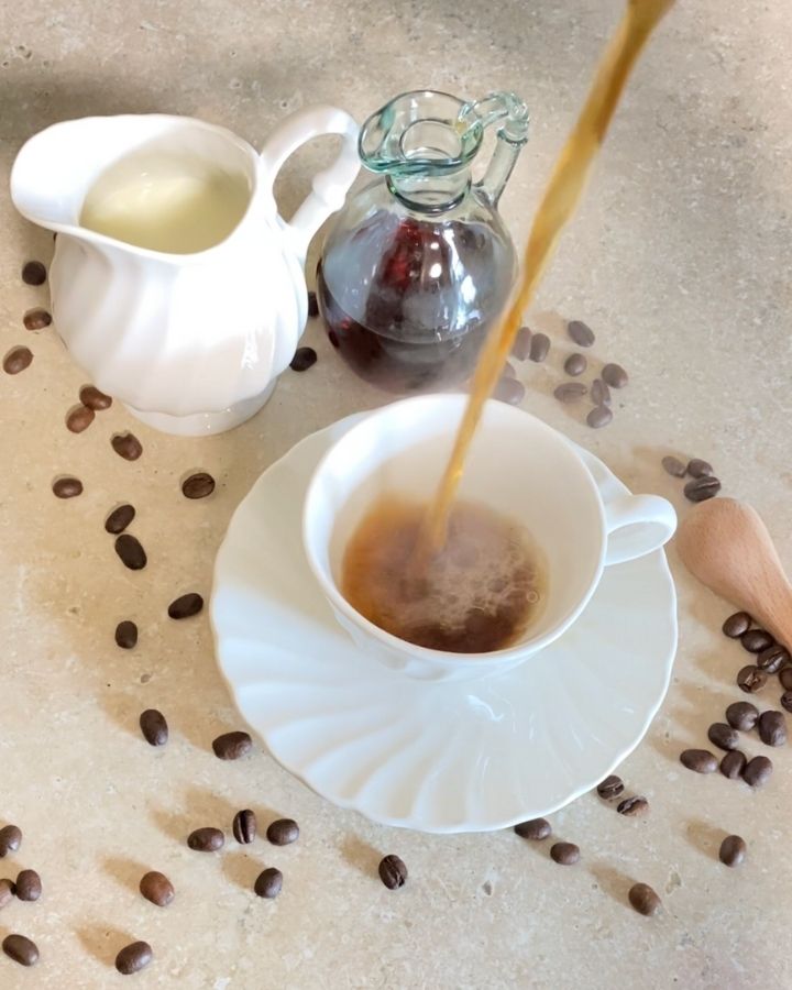 Pouring coffee into a white coffee cup with a syrup jar and cream container beside the cup.